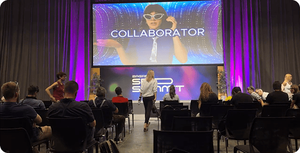 Members at a conference in Zagreb, seated in chairs and facing a large screen displaying the Collaborator promo video.